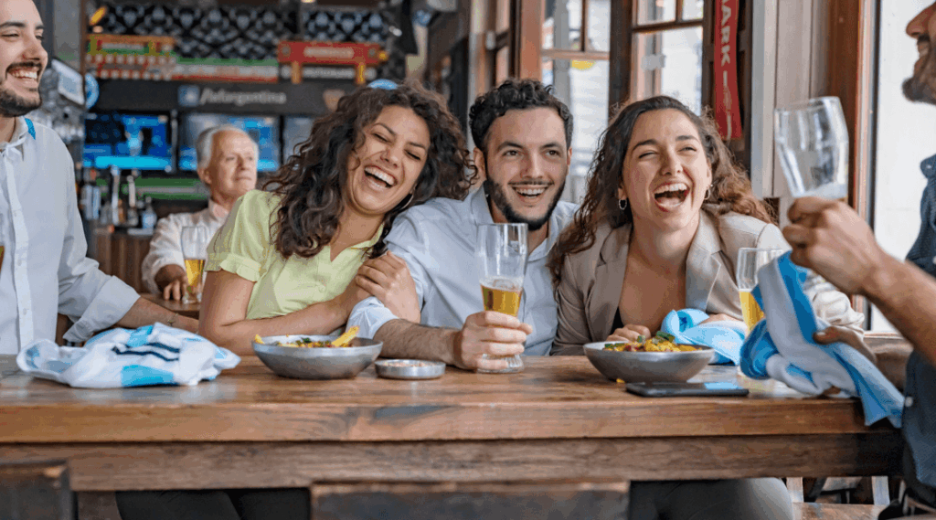 Friends sitting at a bar laughing and holding drinks while watching a sports game on television