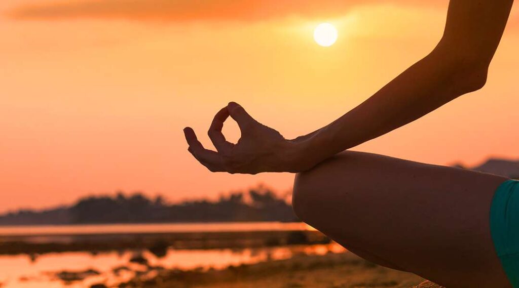 Person sitting outdoors at sunset practicing meditation with hands resting on knees near a body of water
