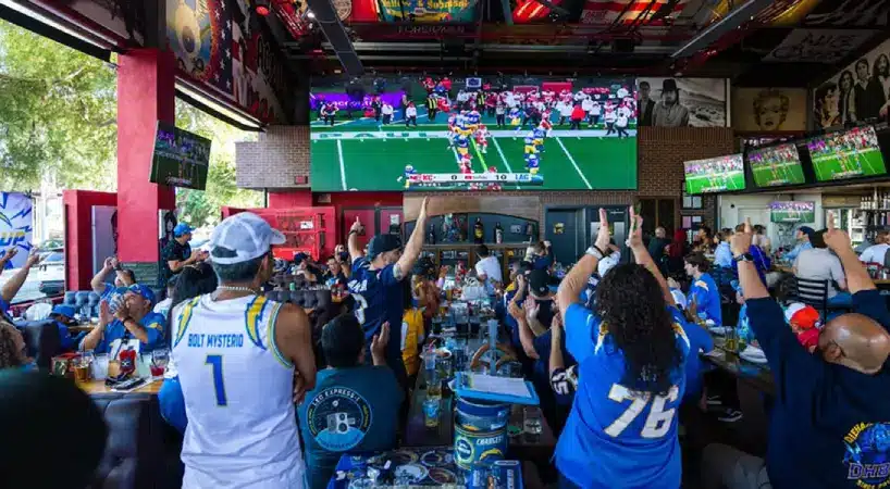 A packed sports bar erupts in celebration as fans in Los Angeles Chargers jerseys and hats raise their arms and cheer. A large screen above the bar shows a football game in progress, with more TVs mounted along the walls.