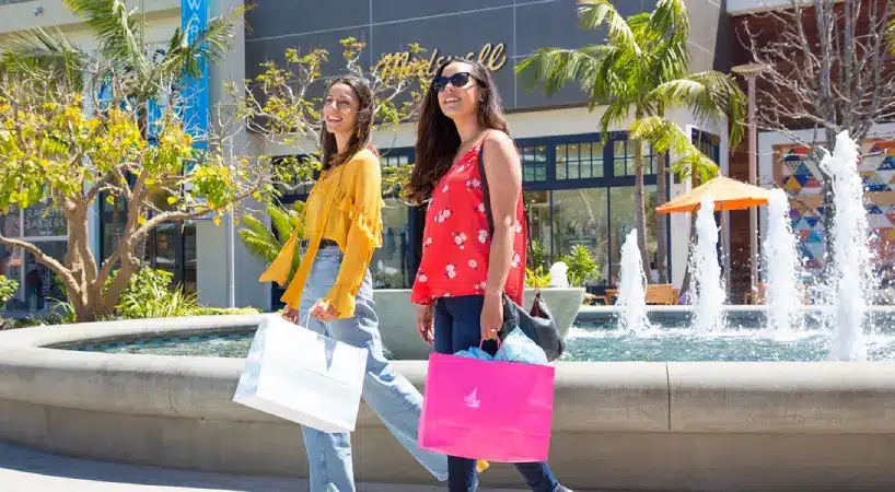 Two smiling women stroll past a fountain at an open-air shopping center on a sunny day, carrying white and hot-pink shopping bags. Palm trees and a Madewell storefront line the plaza behind them.