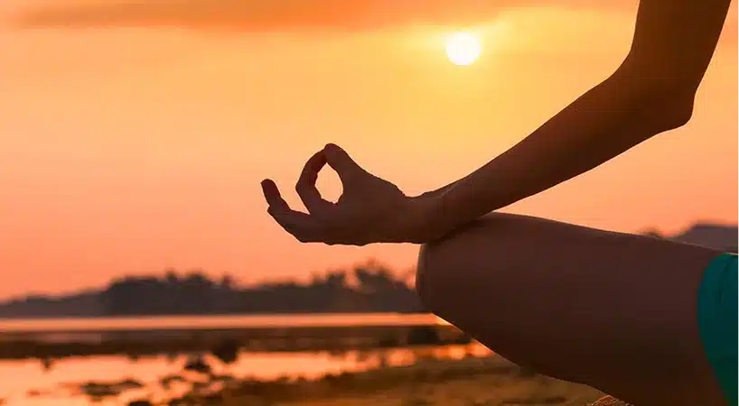Close-up silhouette of a person's hand resting on their knee in a meditation mudra, thumb and index finger touching, as the sun sets over a calm shoreline in shades of orange and pink.