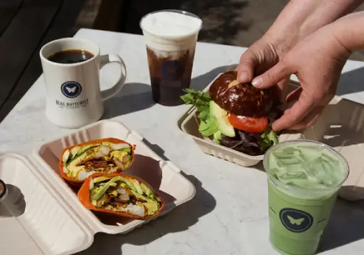 Overhead view of a café spread on a white marble table: a breakfast burrito cut in half in a takeout container, a burger with avocado, tomato, and greens on a dark brioche bun, a mug of black coffee, an iced coffee with cold foam, and an iced matcha latte. The mug and matcha cup feature a butterfly logo.