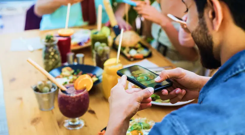 Over-the-shoulder view of a man photographing his meal with a smartphone at a sunny outdoor table. The table is filled with colorful dishes, salads, a burger, fruity drinks garnished with orange slices, and iced beverages with straws, while friends around him reach for food and use their phones.